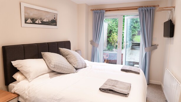 A neatly made bed with pillows, a bedside table, a framed picture on the wall, and a window with curtains. - A Stay On The Brae (Edinburgh)