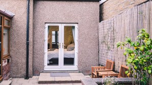 A small house with a sliding glass door leading to a bedroom with a bed and a window. There is a patio area with a wooden table and chairs, and a small garden with plants. - A Stay On The Brae (Edinburgh)