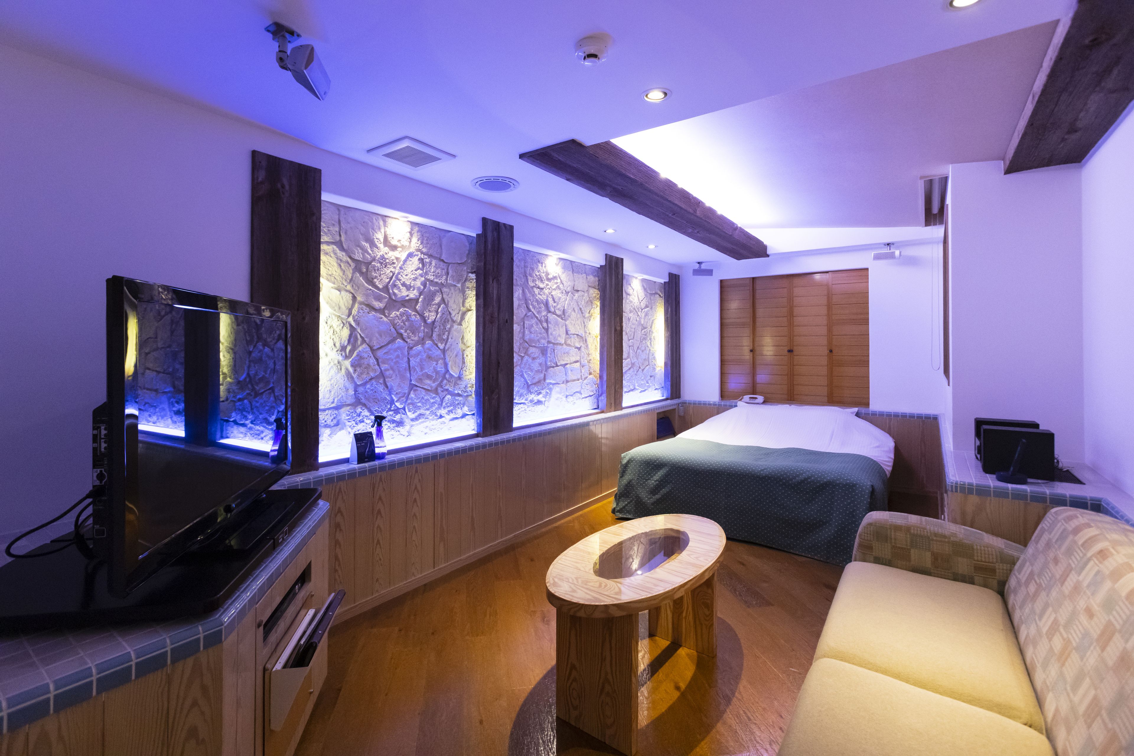 Una habitación de hotel moderna con una cama grande, un televisor de pantalla plana, una pared de piedra, vigas de madera y una pequeña mesa redonda.