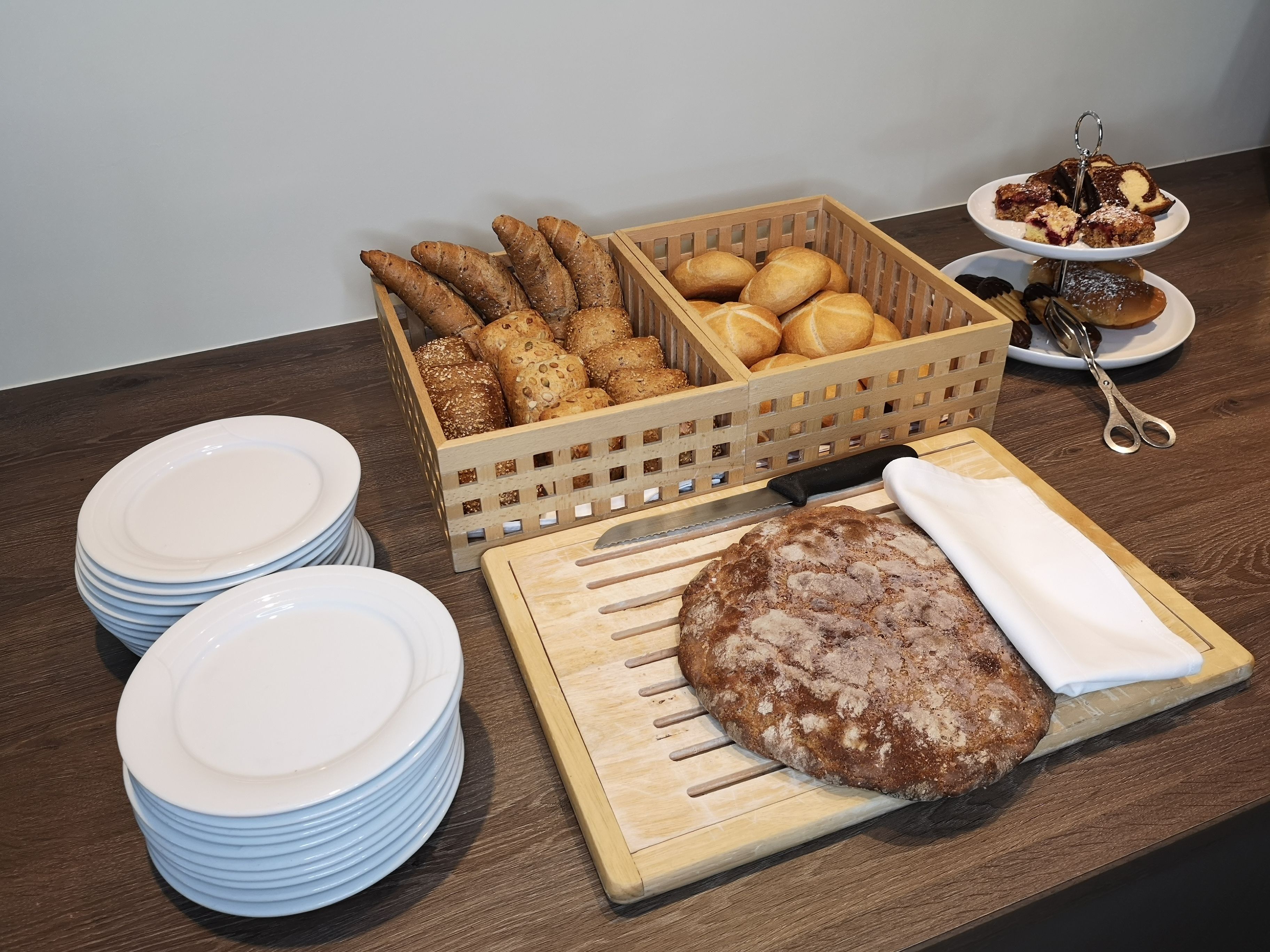 Ein Holztisch mit Stapeln weißer Teller, ein Holzschneidebrett mit Brot, ein Korb mit verschiedenen Brotsorten und ein Dessertteller mit Kuchen und einem Löffel.