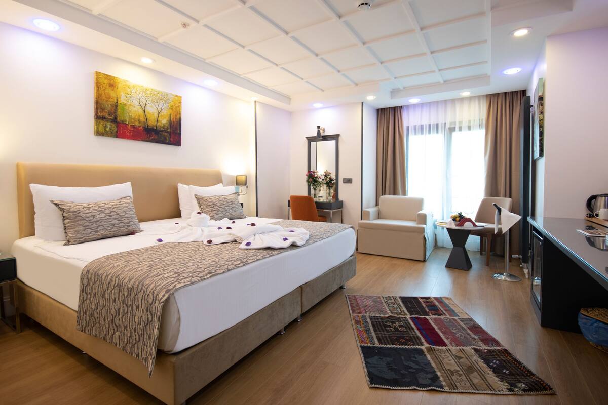 A hotel room with a large bed, a desk, a chair, a sofa, a mirror, and a painting on the wall.