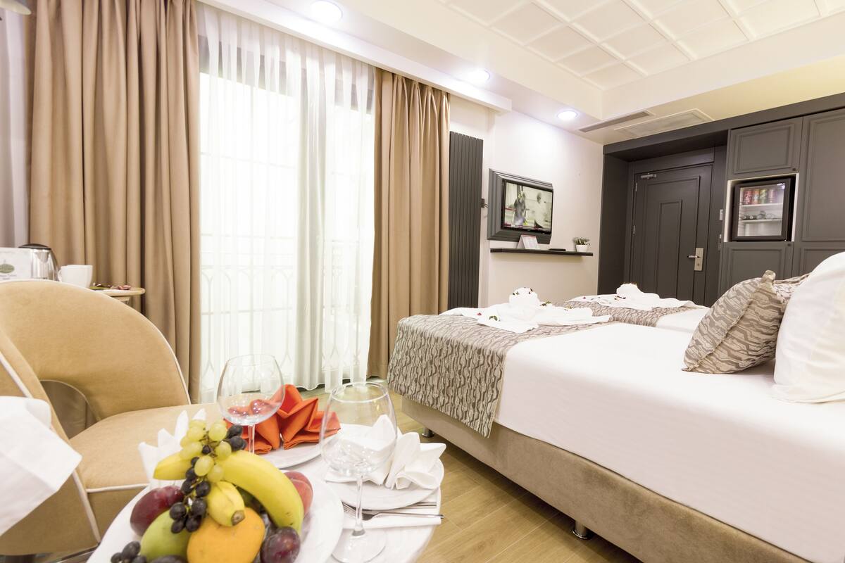 A hotel room with a bed, a dining table with fruit and wine, a TV, and a minibar.