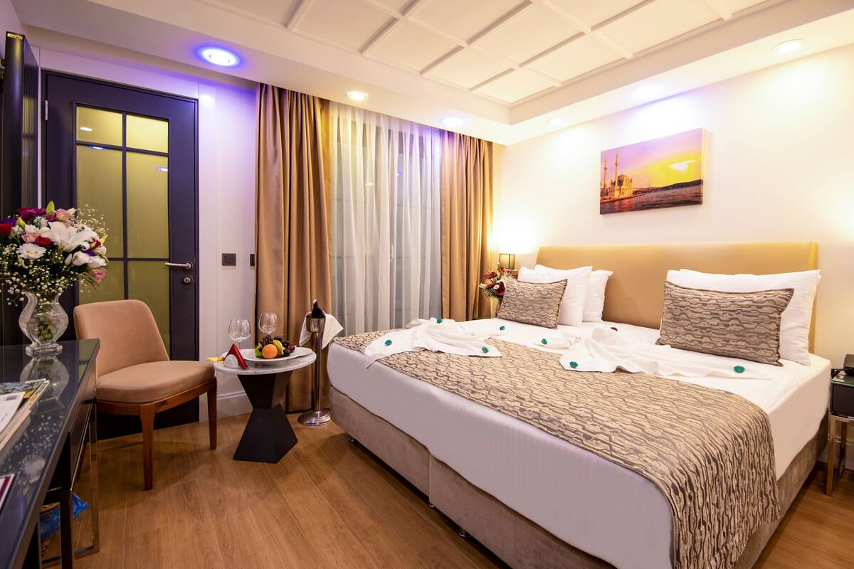 A hotel room with a large bed, a desk with a chair, a small table with a vase of flowers, and a painting on the wall.