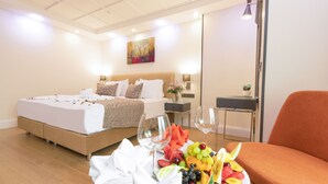 Una habitació d'hotel amb un llit, una taula de menjador amb fruita i gots de vi, i una tauleta de nit amb una làmpada.