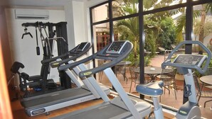 A gym with treadmills and an elliptical machine, large windows, and outdoor seating visible through the windows. - Golden Valley Hotel Enugu (Nsukka)