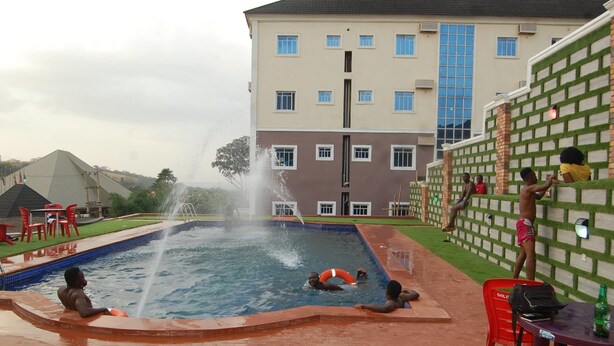 A swimming pool with a water fountain, surrounded by a brick wall and a building with multiple windows. - Golden Valley Hotel Enugu (Nsukka)
