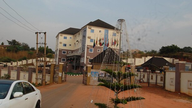 A gated entrance to a hotel with multiple flags, a tall structure, and a car parked in front. - Golden Valley Hotel Enugu (Nsukka)
