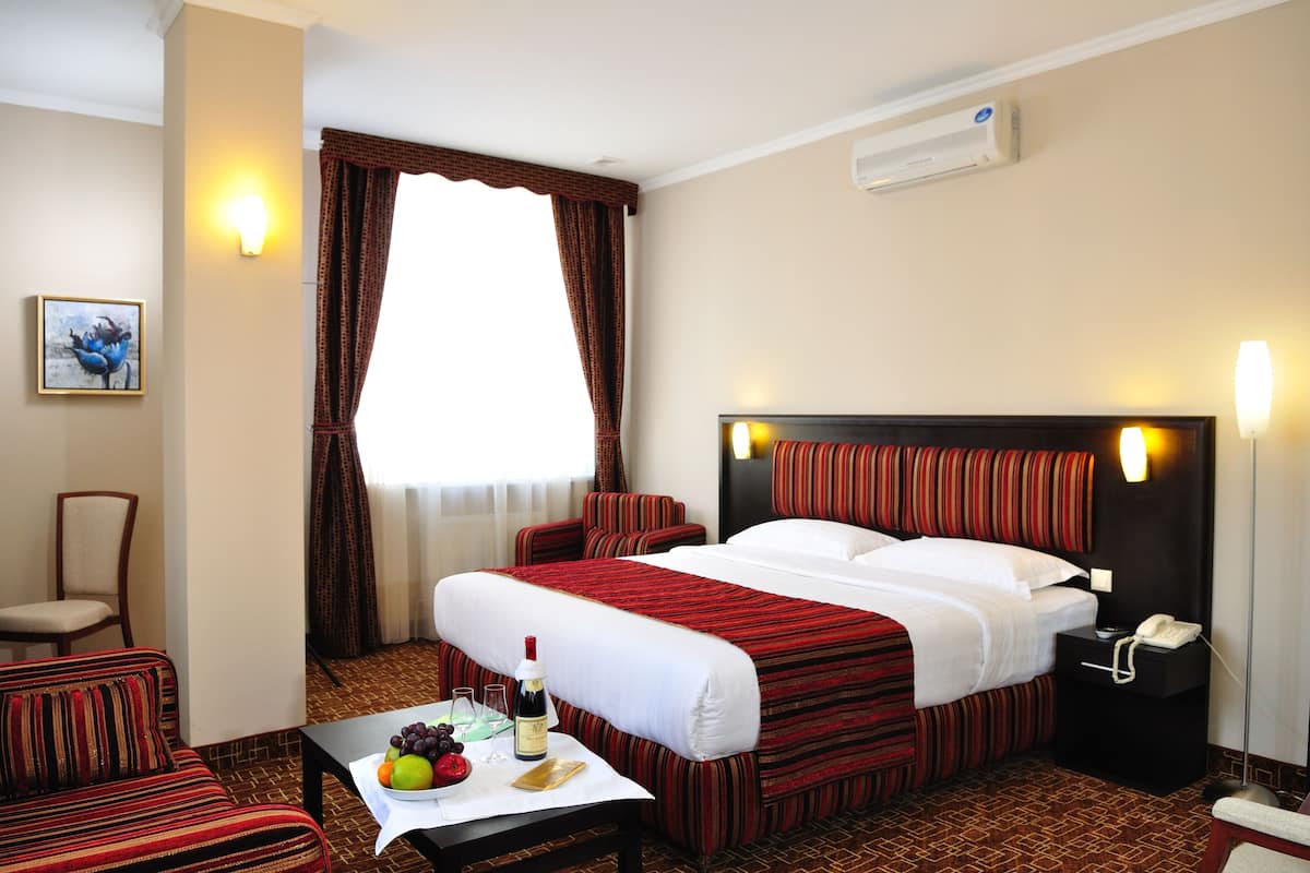 A hotel room with a large bed, a small table with fruits and a bottle, a chair, a nightstand with a phone, and a wall-mounted air conditioner.