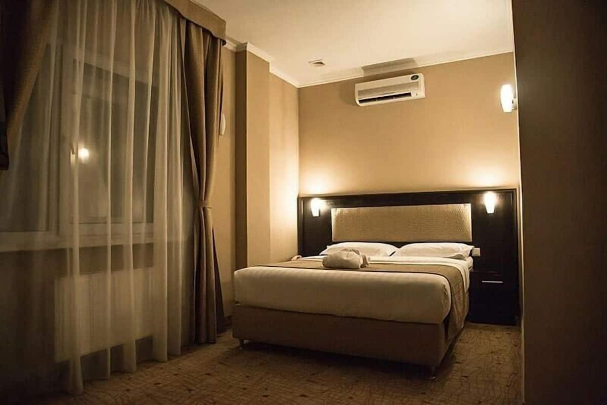 A hotel room with a bed, curtains, an air conditioning unit, and a nightstand.