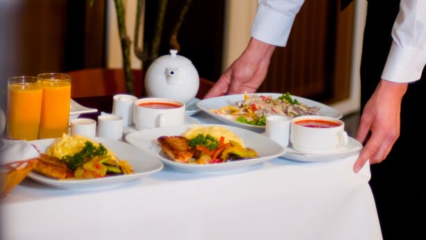 A waiter serving breakfast at a table with plates of food, a teapot, and two glasses of orange juice.