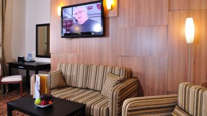 A hotel room with a television mounted on the wall, a striped sofa, a small table with a fruit bowl, and a desk with a chair.