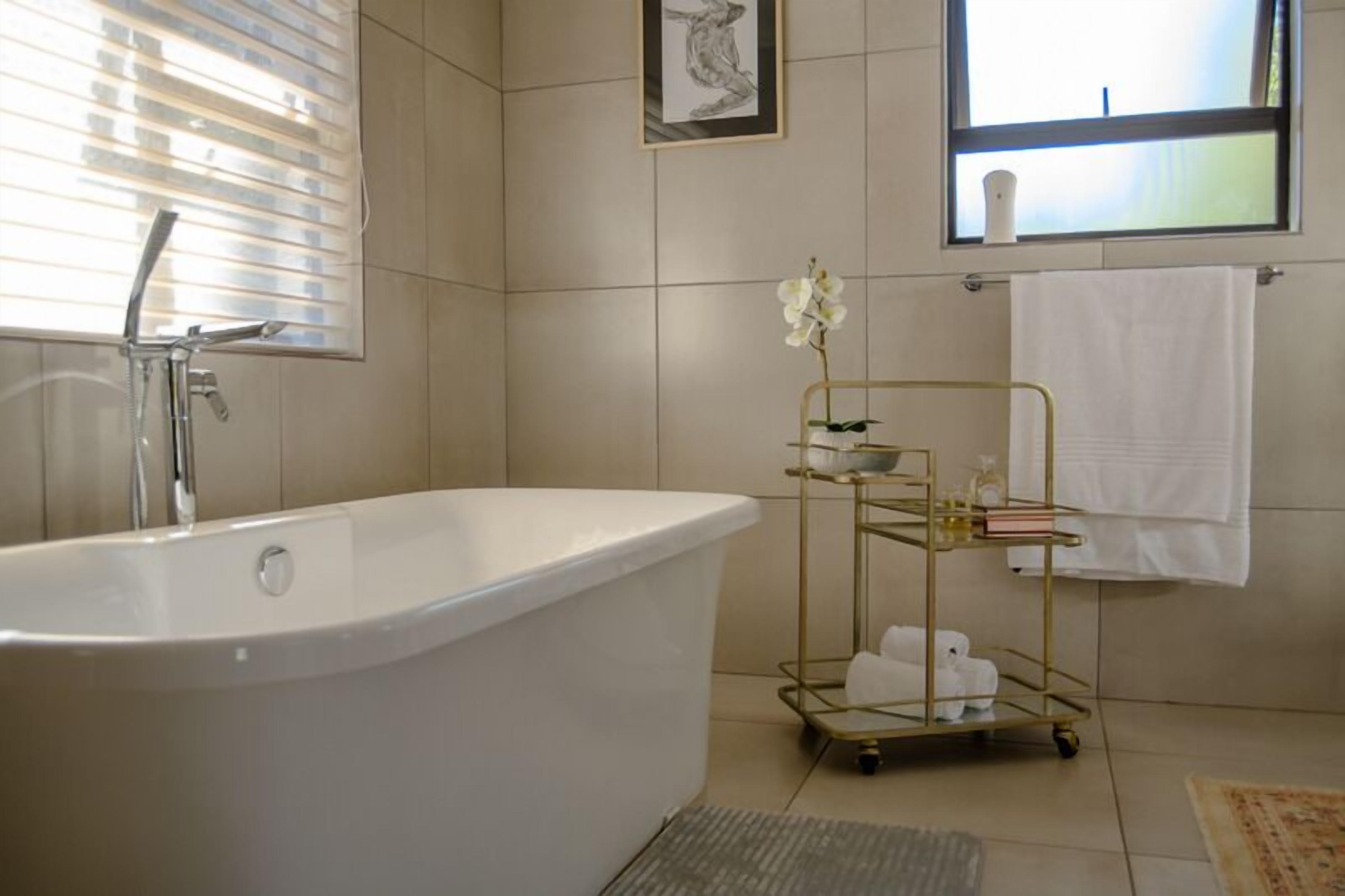 Une salle de bain moderne avec une baignoire sur pieds, un chariot doré contenant des articles de toilette, une œuvre d’art encadrée au mur et une fenêtre avec des stores.
