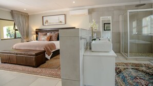 A modern bedroom with a large bed, a bedside table, a nightstand, a bathroom with a shower, and a glass-enclosed walk-in closet. - The Reserve Boutique Hotel (Mbombela)