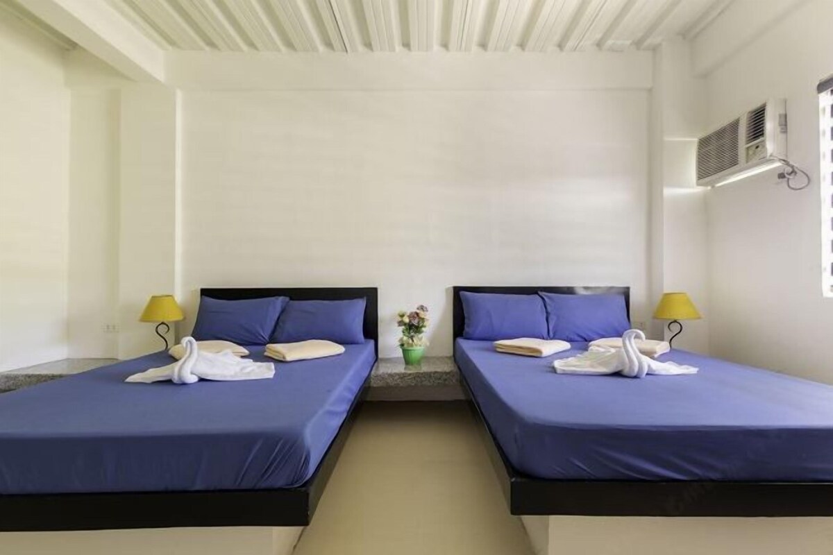 A hotel room with two beds, each with a blue bedspread and pillows, a small table with a lamp, and a potted plant.