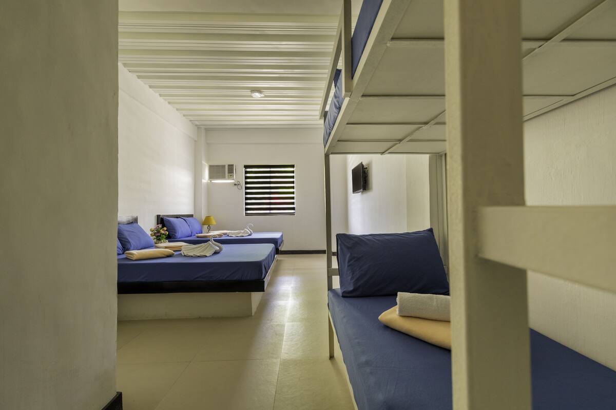 A room with bunk beds, a small window with blinds, a wall-mounted TV, and a bedside table with a lamp.