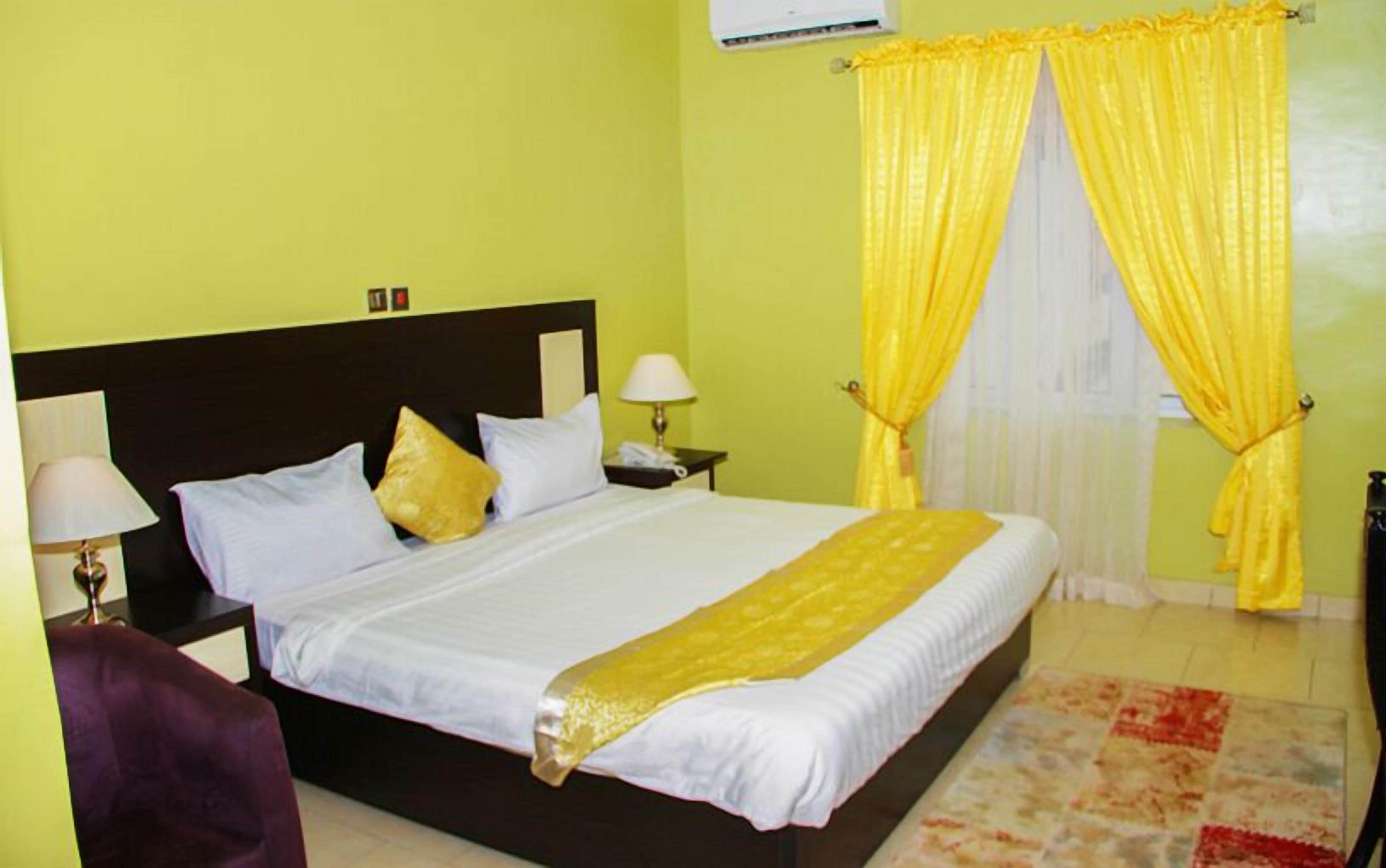 A hotel room with a bed, two bedside lamps, a nightstand, a purple chair, and yellow curtains.