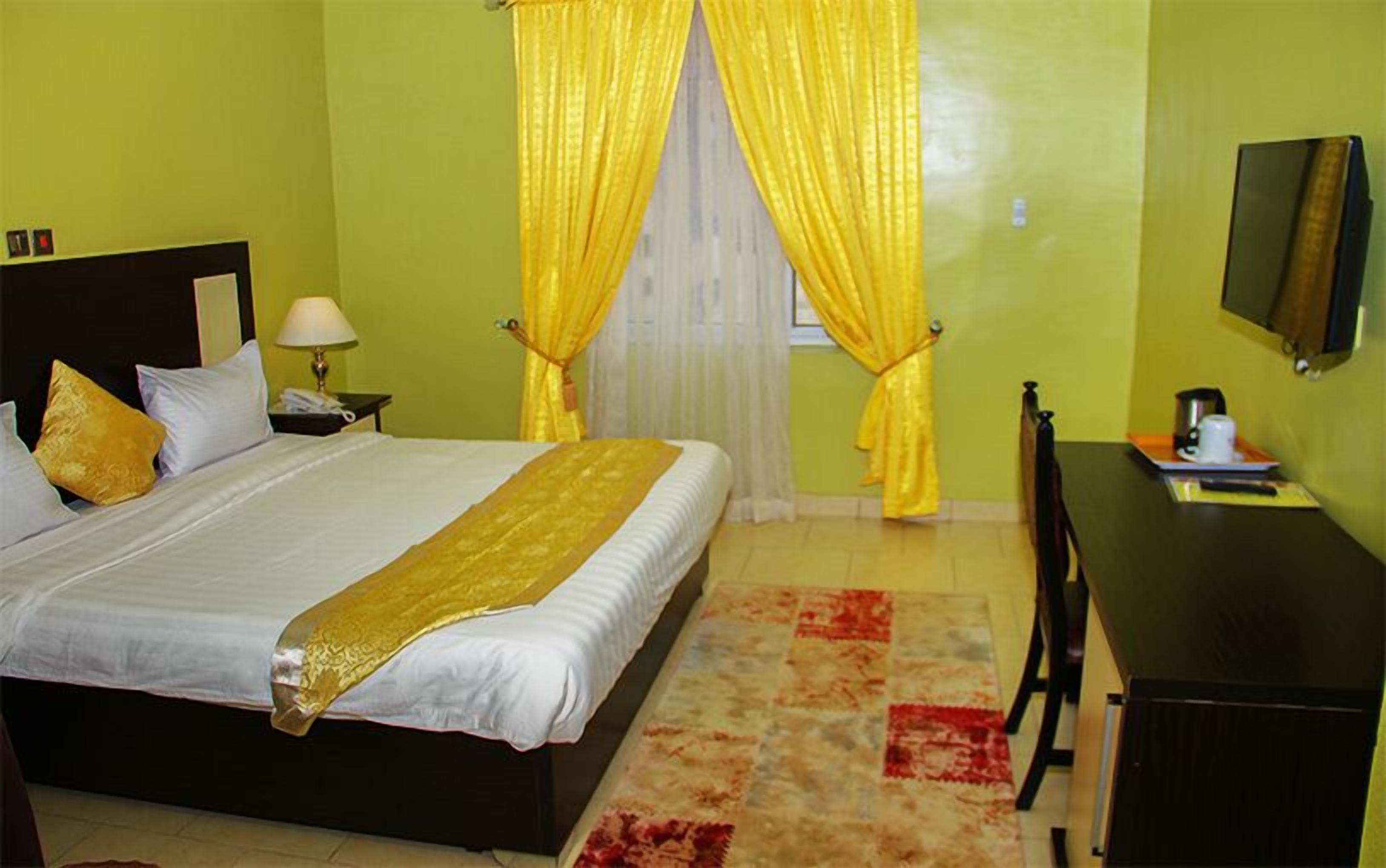 A hotel room with a bed, a desk, a chair, a TV, and a window with yellow curtains.