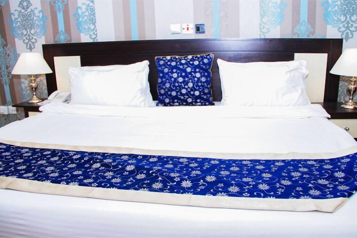 A neatly made bed with white linens, a blue floral bedspread, and two matching pillows. There are two bedside lamps and a small table with a phone and a decorative item.