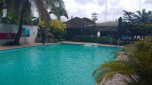 A swimming pool with a clear blue water, surrounded by palm trees and a building with a tiled roof. - Psalm Hotel (Enugu)