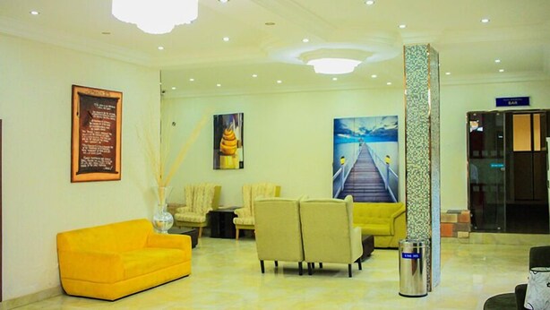 A hotel lobby with a yellow sofa, two armchairs, a coffee table, a large painting of a bridge, and a pillar with a patterned design. - Psalm Hotel (Enugu)