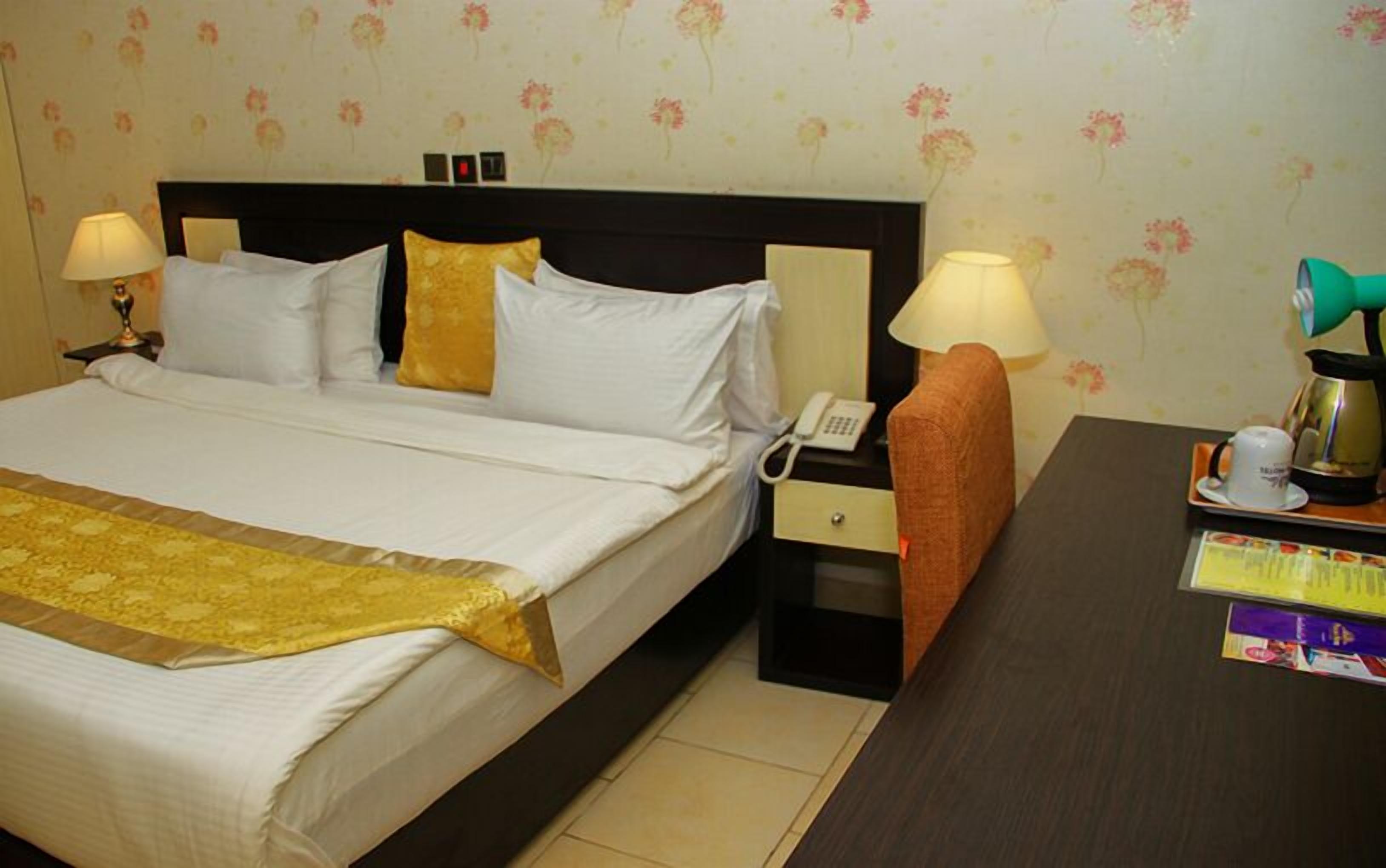 A hotel room with a large bed, two bedside lamps, a nightstand with a phone, a small table with a teapot and cups, and a floral wallpaper.