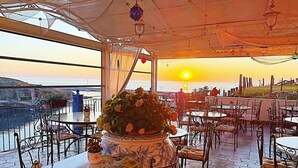 Un área de comedor al aire libre con mesas y sillas, un jarrón grande con flores y vistas al atardecer sobre el mar.