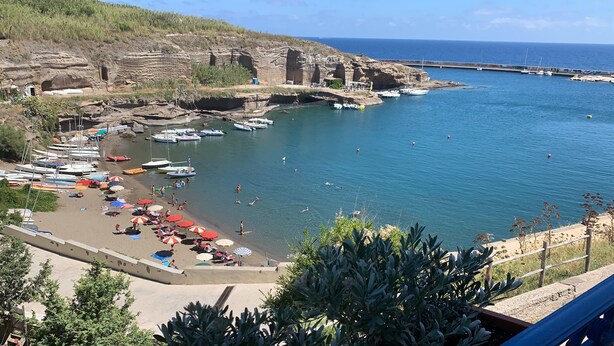 Una zona costera con barcos amarrados, una playa arenosa y acantilados rocosos.