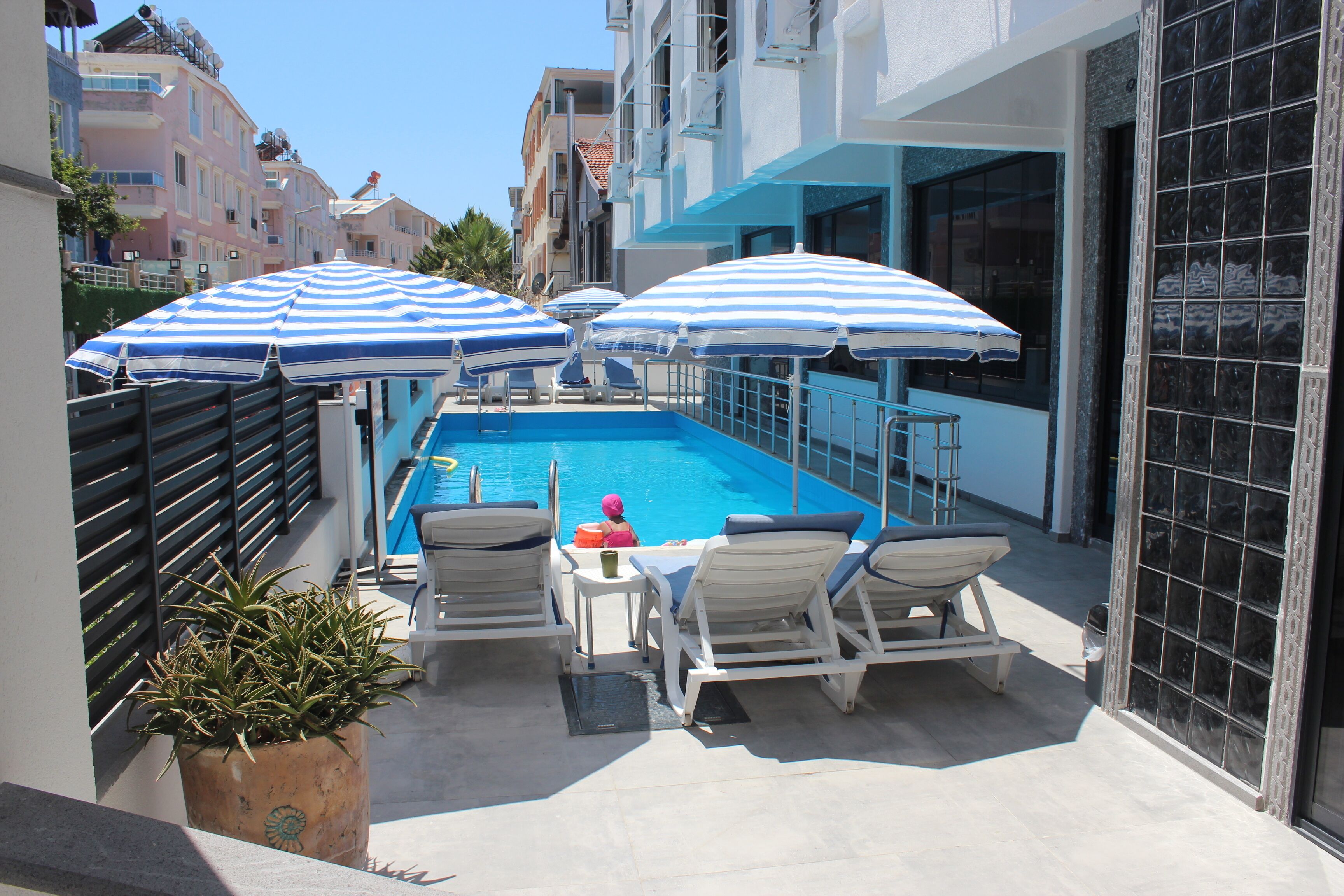 A rooftop pool area with blue and white striped umbrellas, lounge chairs, and a potted plant.