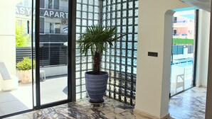 A corner room with a large potted plant, a sliding glass door leading to an outdoor area, and a view of a building with 'APART' signage. - Tasan Apart Otel (Didim)
