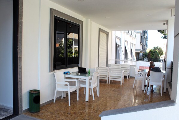 A clean, modern outdoor dining area with white furniture and a glass table, featuring a view of the street and buildings in the background. - Tasan Apart Otel (Didim)