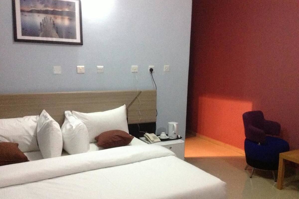 A hotel room with a bed, a nightstand with a phone and a coffee maker, a chair, and a red wall.