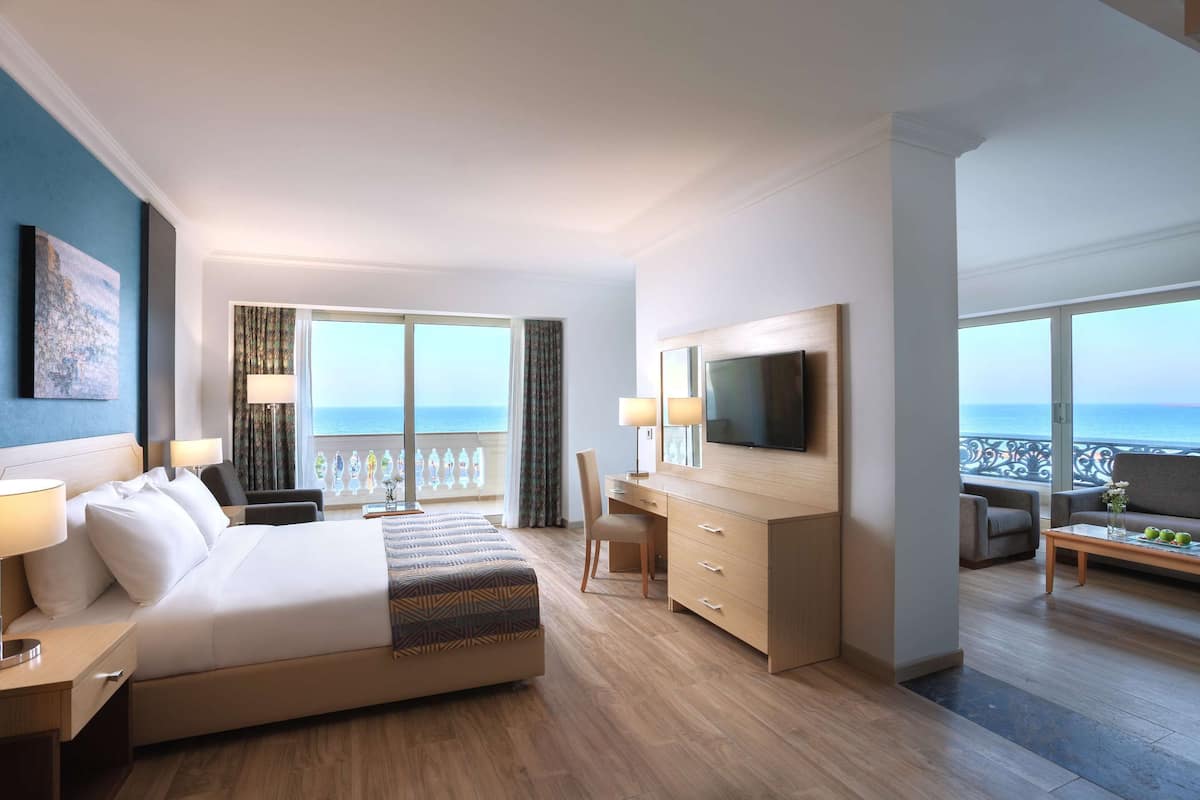 A hotel room with a large bed, a desk, a TV, and a balcony with a view of the sea.