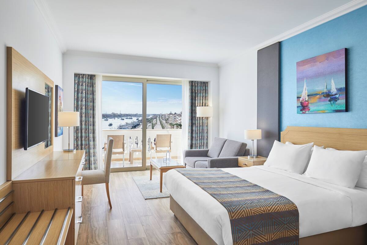 A hotel room with a large bed, a desk, a chair, a TV, and a balcony with a city view.