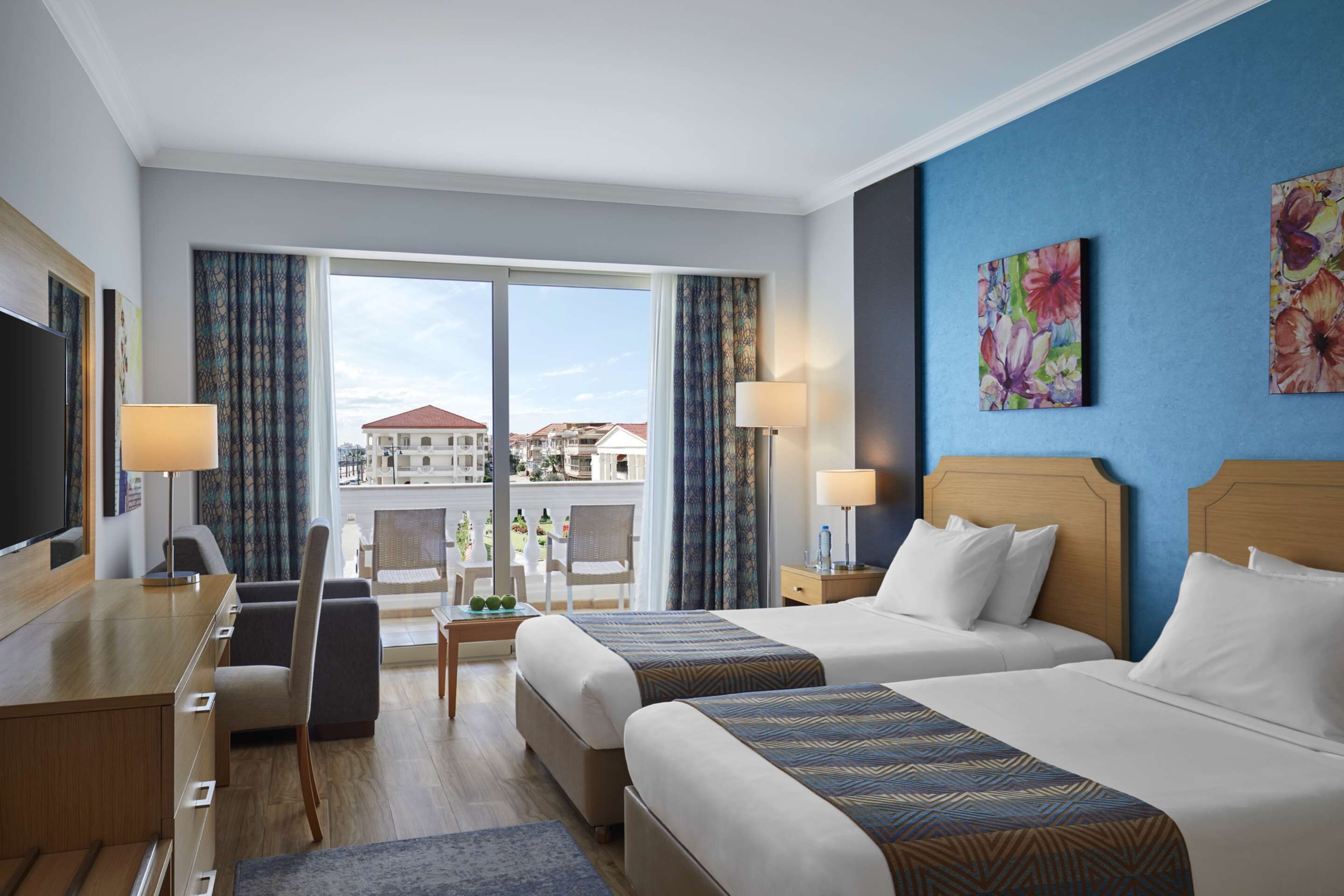 Habitación de hotel con dos camas, un escritorio, un televisor y un balcón con vistas a edificios.