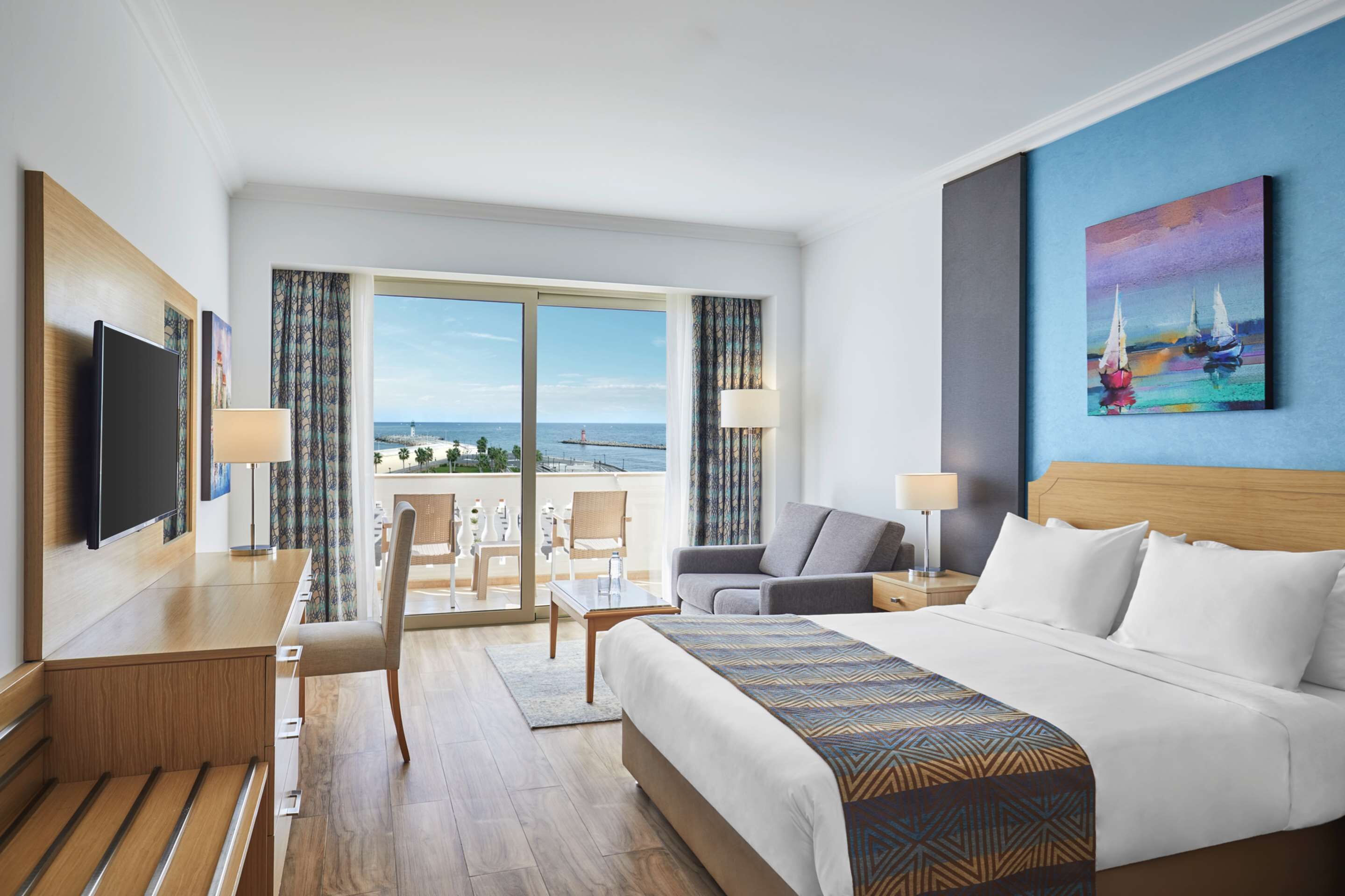 A hotel room with a large bed, a desk with a flat-screen TV, a chair, a sofa, and a view of the sea through large windows.