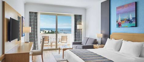 A hotel room with a large bed, a desk with a flat-screen TV, a chair, a sofa, and a view of the sea through large windows.