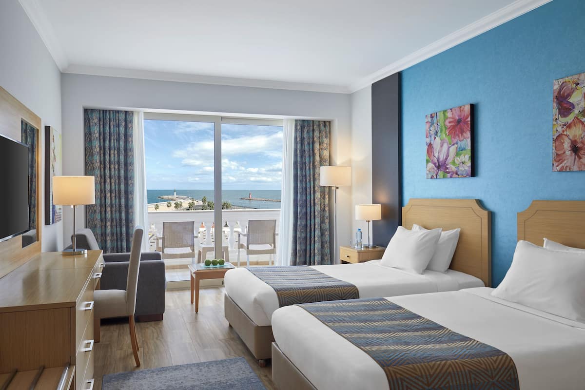 A hotel room with two beds, a desk, a TV, and a balcony with a view of the sea.