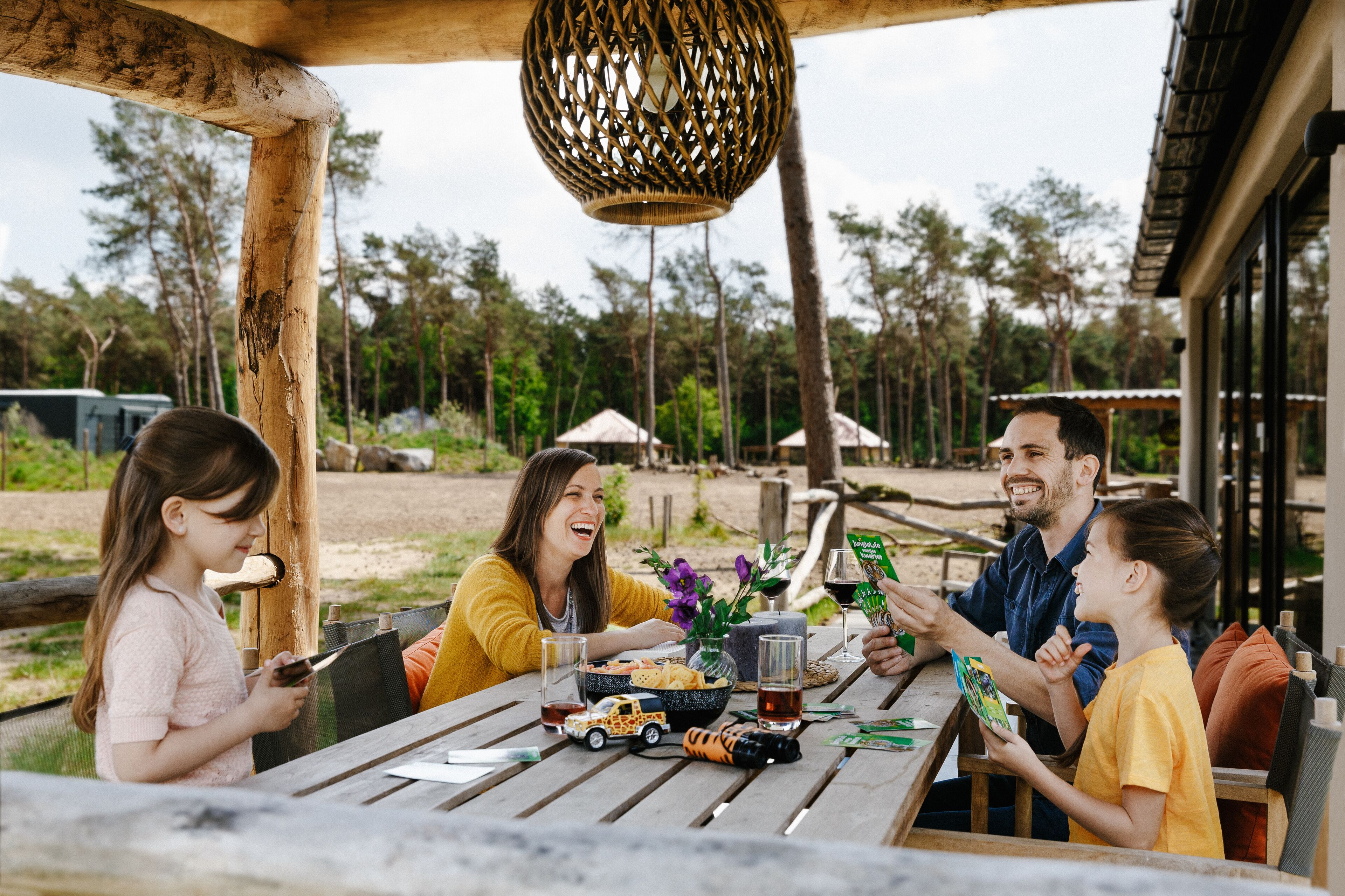 Een houten tafel met een hanglamp van gevlochten materiaal erboven, omringd door mensen die zitten aan een picknicktafel met eten en drinken.