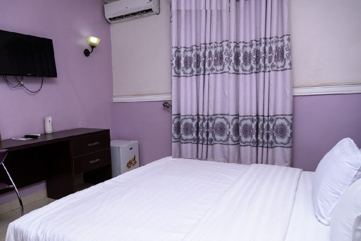 A hotel room with a bed, a desk, a chair, a lamp, and an air conditioning unit.
