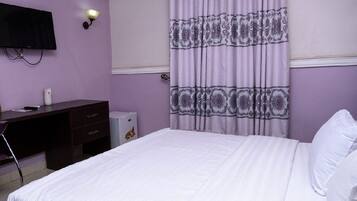 A hotel room with a bed, a desk, a chair, a lamp, and an air conditioning unit.