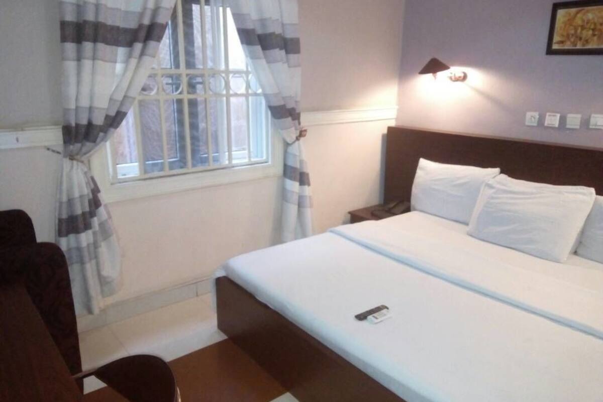 A bedroom with a bed, a nightstand, a lamp, a window with curtains, and a chair.