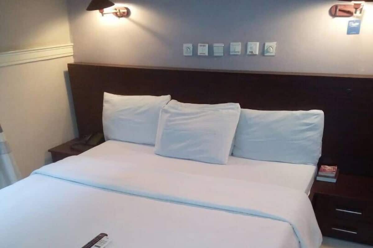 A neatly made bed with white linens, a remote control on the bed, and a nightstand with a lamp and a book beside it.
