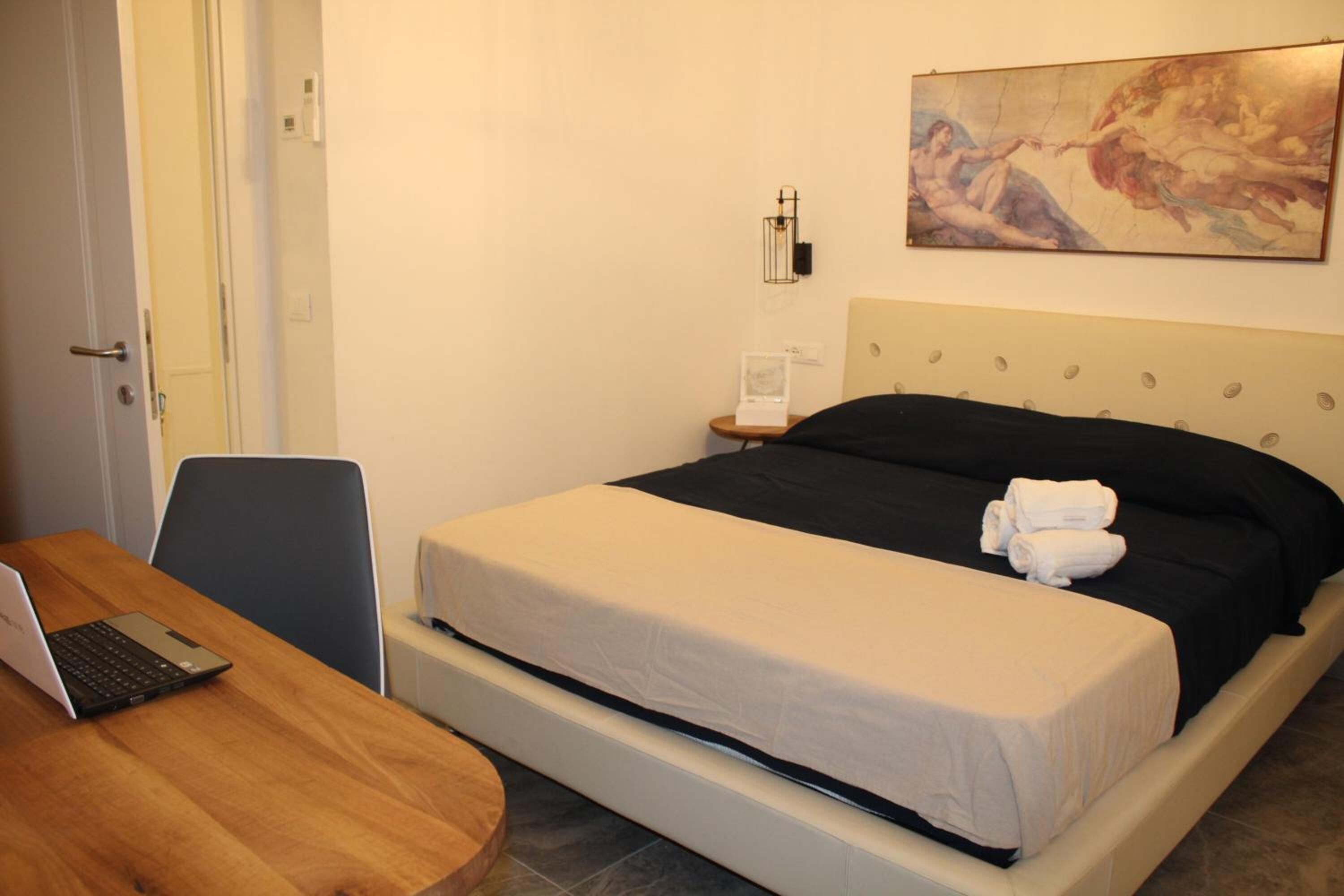 A hotel room with a bed, a desk with a laptop, a nightstand, and a painting on the wall.