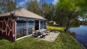A small house with a patio by a lake, featuring a table and benches, surrounded by trees. - Lake Resort Beekse Bergen (Hilvarenbeek)