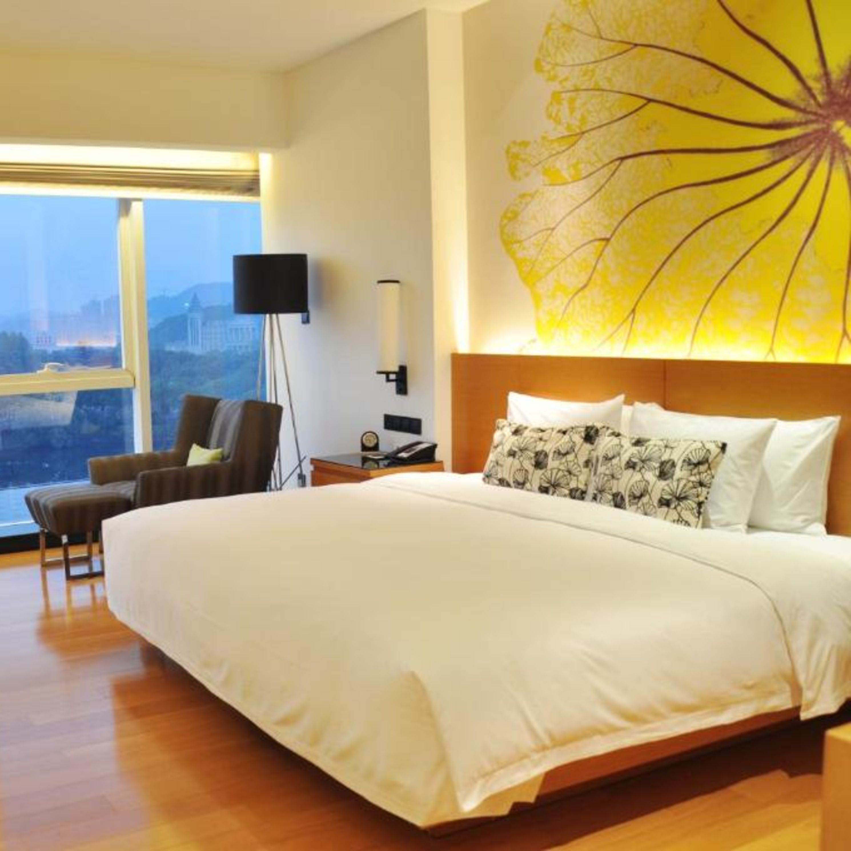 A hotel room with a large bed, a chair, a desk, and a view of the city through a large window.