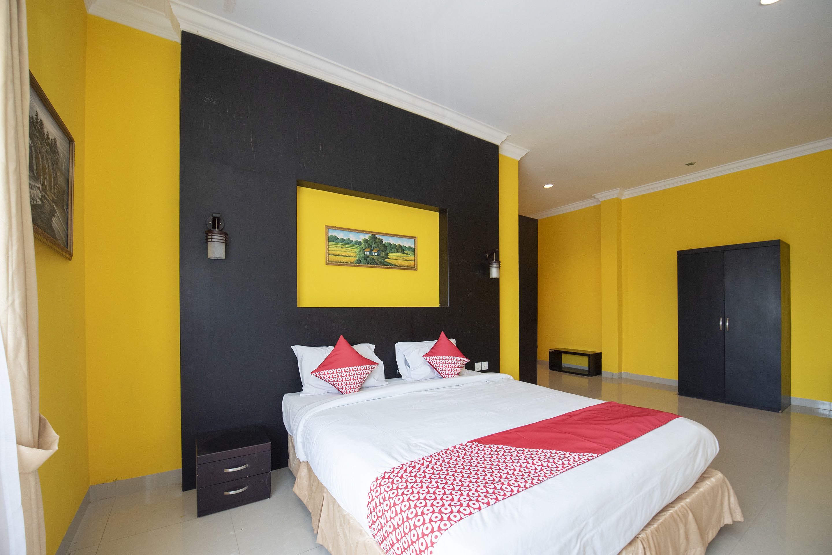 A hotel room with a large bed, a black headboard, a yellow accent wall, and a framed picture above the bed.