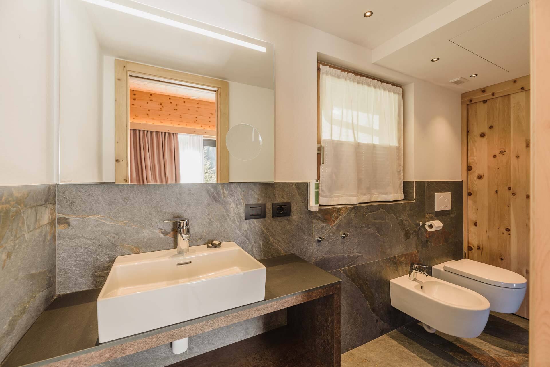 A modern bathroom with a large mirror, two sinks, a toilet, and a wooden door.