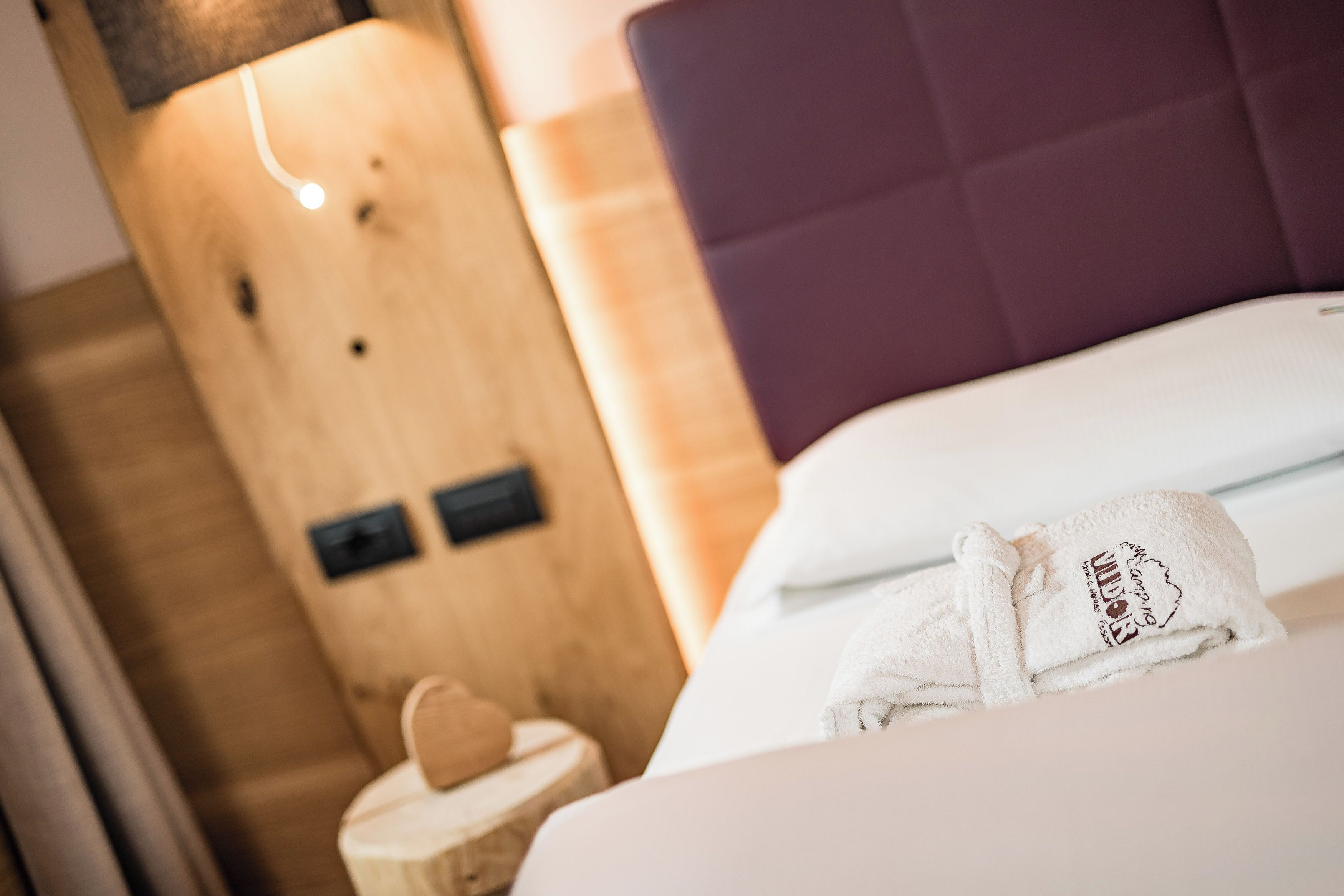 Habitación de hotel con cabecera de madera, cama blanca con toalla blanca y un taburete de madera pequeño con un objeto con forma de corazón.
