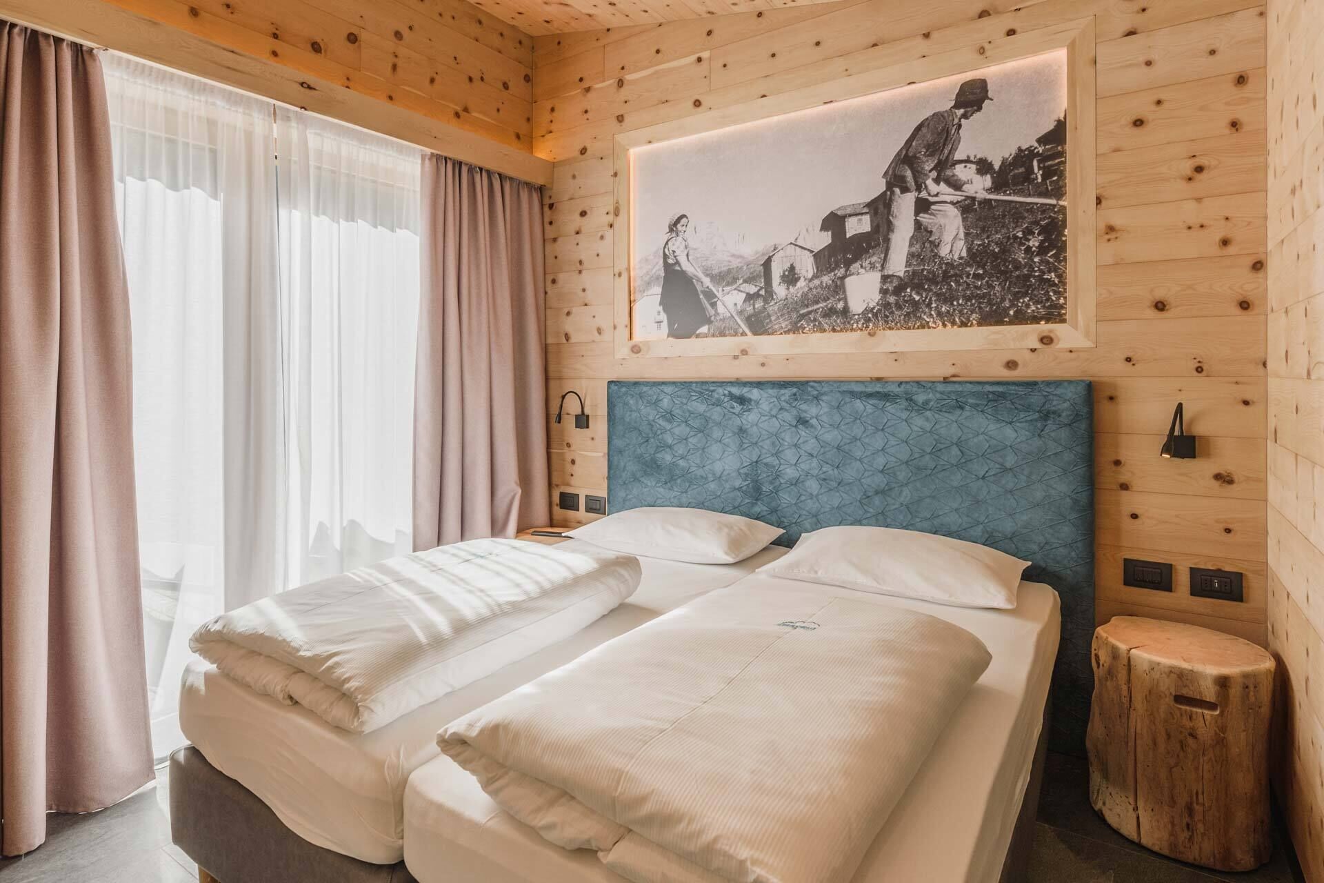 A double bed with a blue headboard, a wooden wall with a framed picture, and a window with sheer curtains.