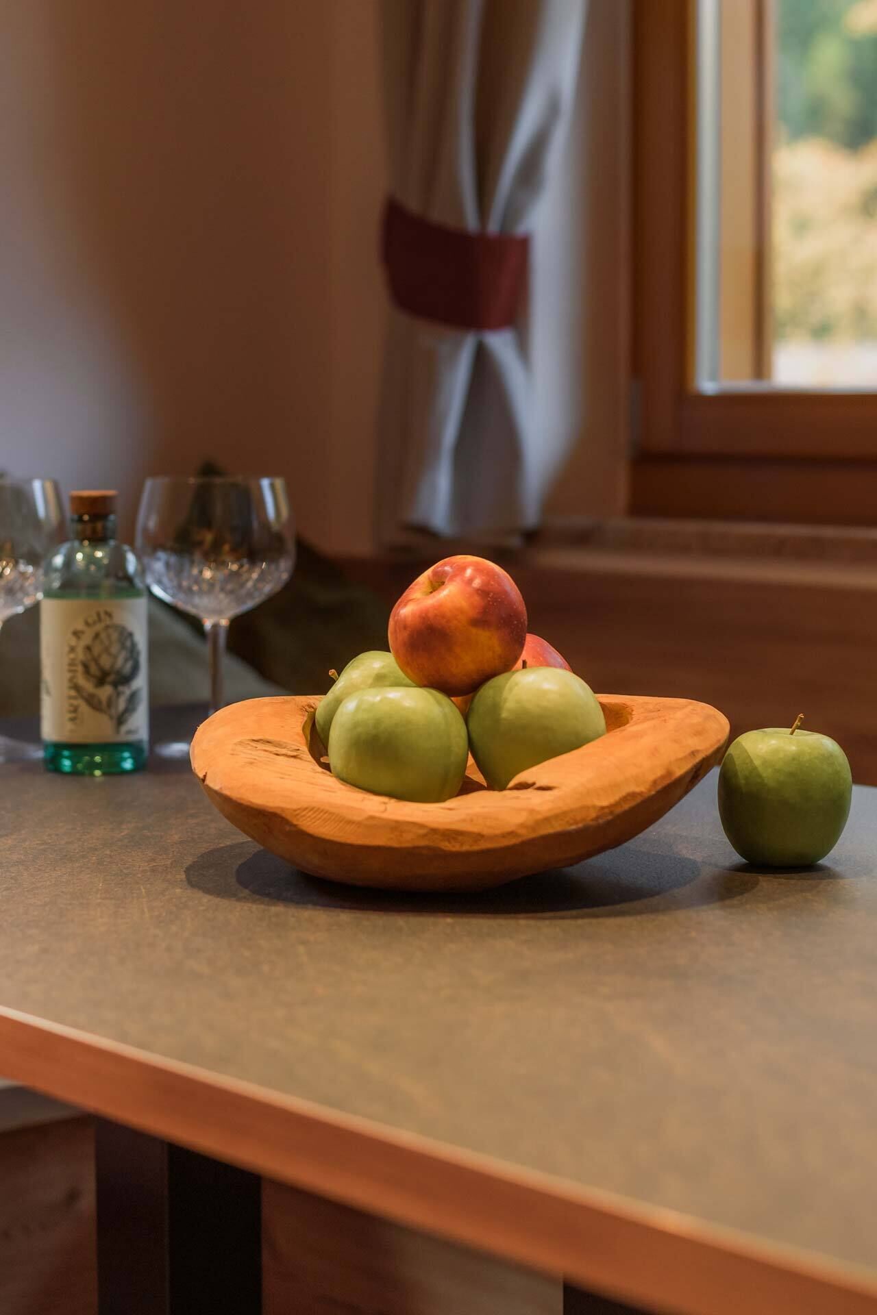A wooden bowl with apples on a table, a bottle of gin, and two glasses.
